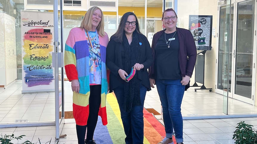 Sally Conning wears a rainbow cardigan and harrier shing and caitlin Grogsby in black standing on a rainbow carpet 