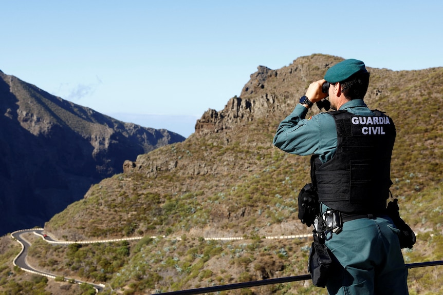 A Guardia Civil officer looks through binoculars to look over rocky mountains and a ravine