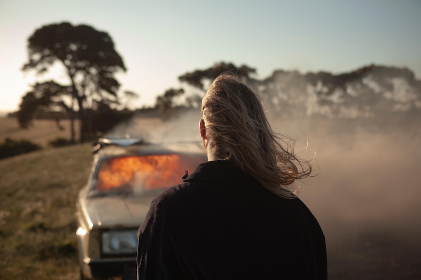 A man with long blond hair stands in front of a burning car.