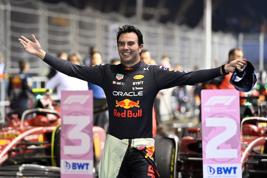 An F1 driver in blue racing suit, stands with his arms out in celebratino, smiling, in front of his car.
