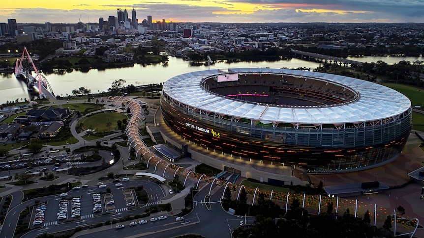 An aerial shot above Perth Stadium lit up with the big screen visible and a bridge, the Swan River and Perth CBD in background.