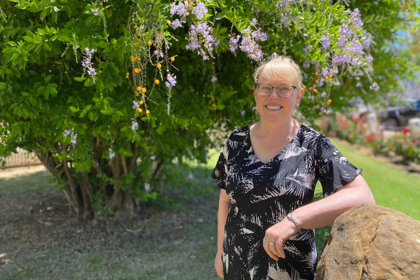 A woman is standing, leaning on a stone, smiling at the camera. There's a blossoming tree behind her.