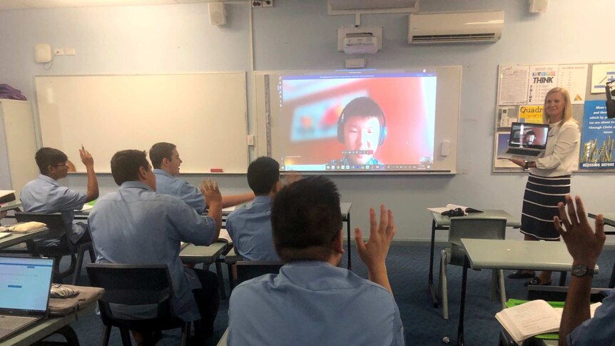 Boys sitting at desks raise their hands, a video of a boy on screen with headphones on is projected onto a whiteboard.