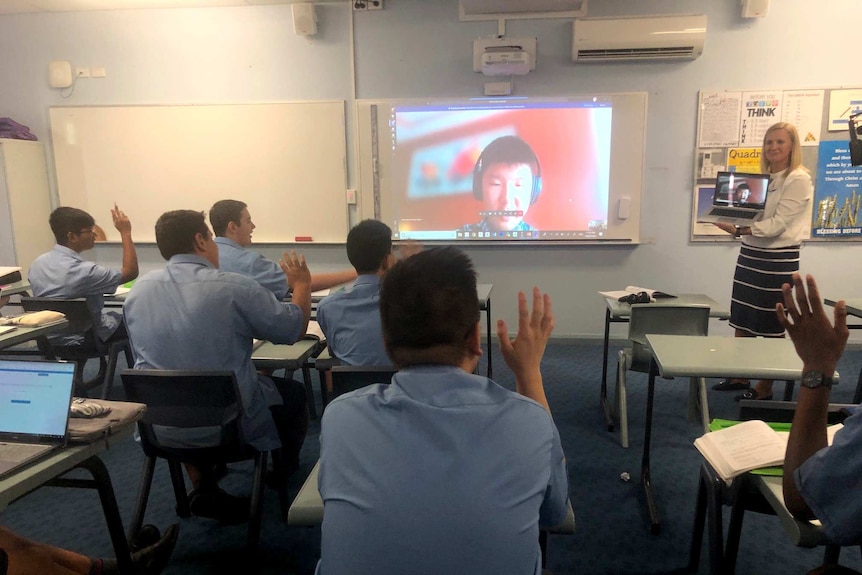 Boys sitting at desks raise their hands, a video of a boy on screen with headphones on is projected onto a whiteboard.