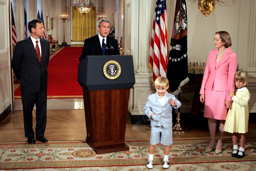 George W Bush stands behind a lectern in the White House with John Roberts to his right, and a woman and two kids to his left 