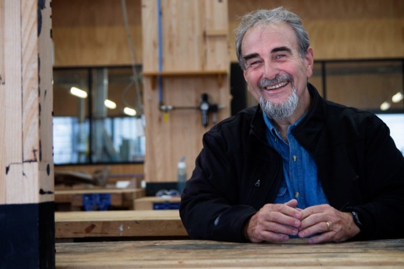 A man with silver hair and beard sits at a timber work bench and smiles
