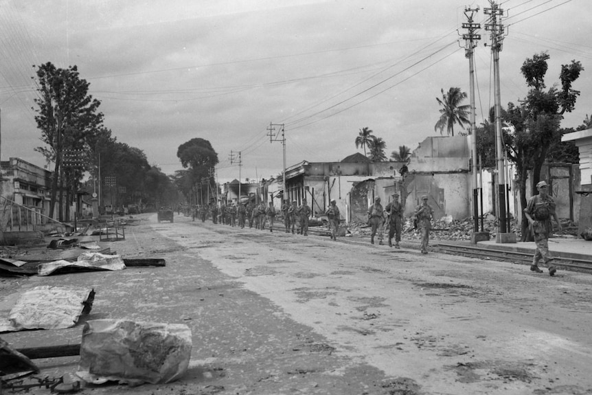 A black-and-white photo of Dutch soldiers walking along a desolate city street, with debris on the ground