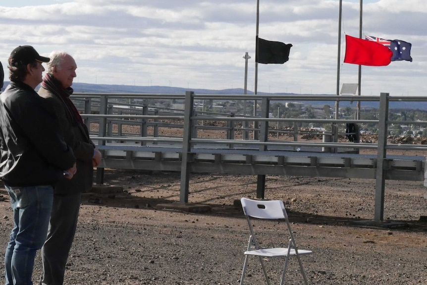 Two men stand near flags, the Australian flag and a black and a red flag, flying at half mast