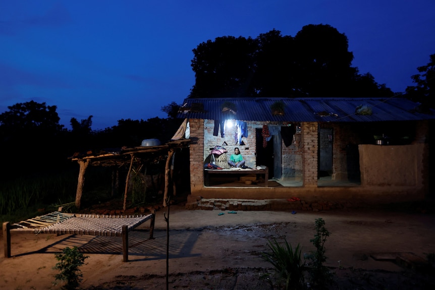 A woman sits on the porch of a brick house at dusk. A side roof and a bed-like structure are in the dirt yard 