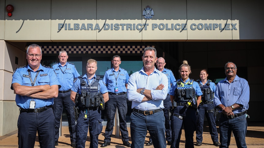 A group of police officers and two politicians stand outside a police station, looking at the camera.