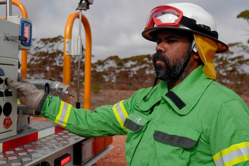 A bearded Indigenous man in firefighting gear stands at the back of a truck in the outback.