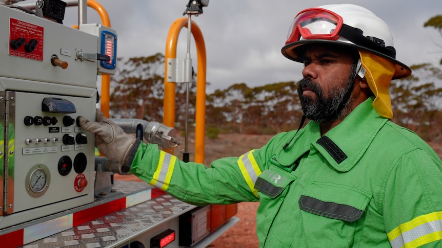 A bearded Indigenous man in firefighting gear stands at the back of a truck in the outback.