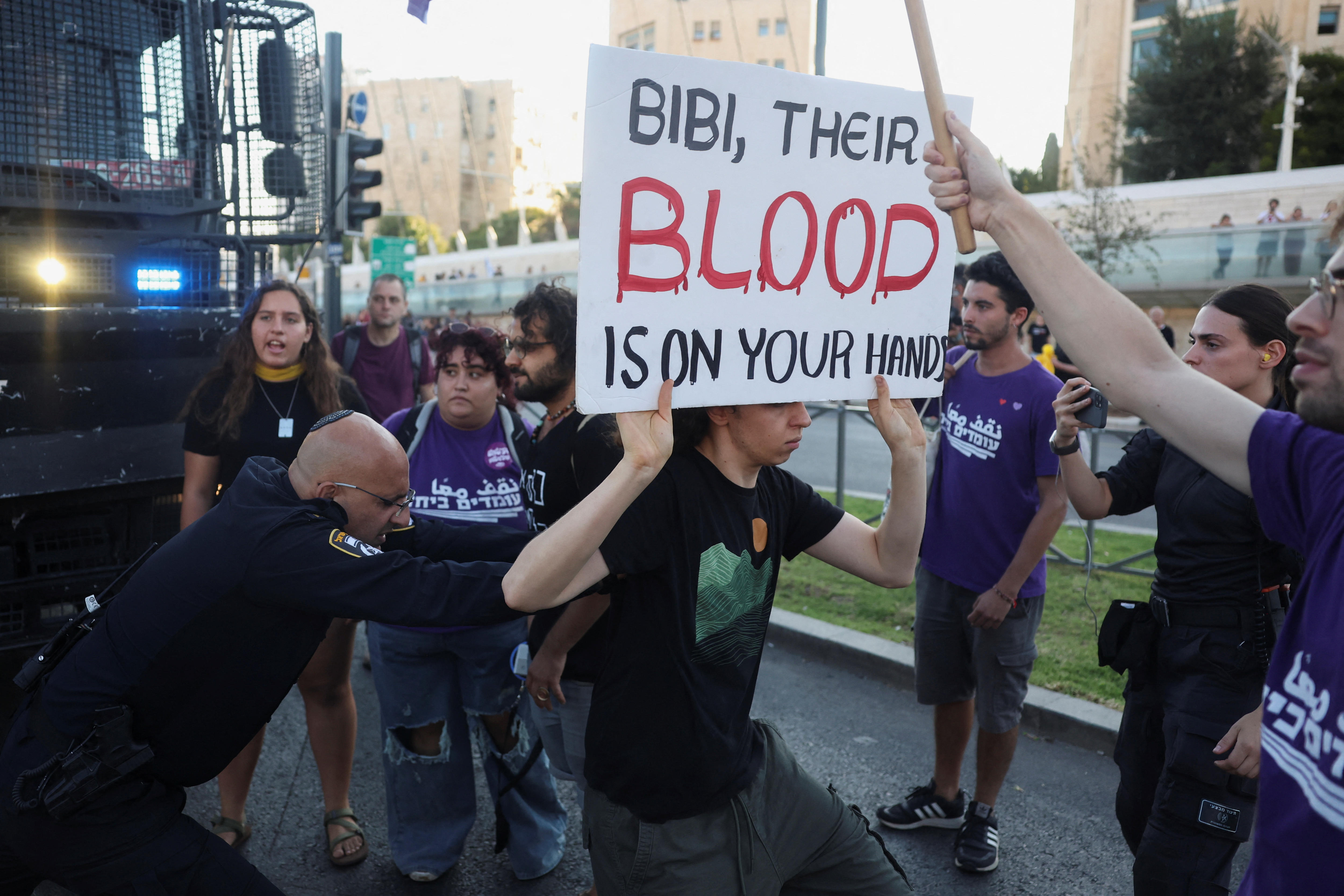 A man holding a sign reading "Bibi, Their blood is on your hands". while being pushed by a security guard surrounded by others