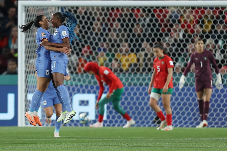 Two soccer players in blue jump in celebration in front of a soccer goal net and players from another team in red.