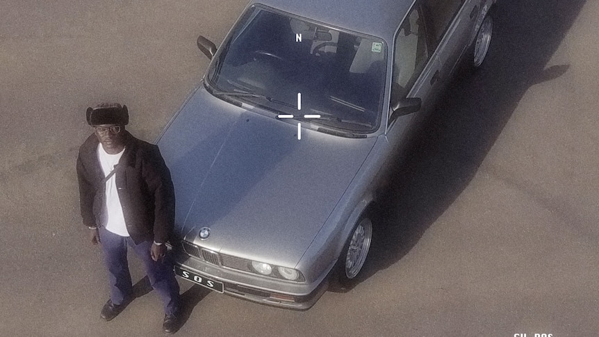 Aerial photo of Skin On Skin, staring upwards & standing next to a silver BMW. He wears a black hat, brown jacket & jeans