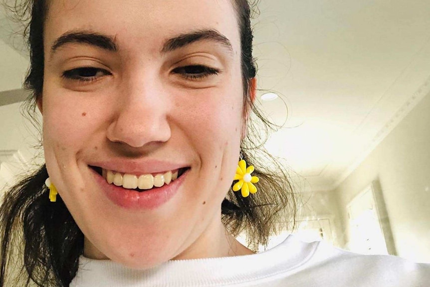 Hannah Diviney souriante selfie assise à l'intérieur dans une pièce blanche, portant des boucles d'oreilles à fleurs