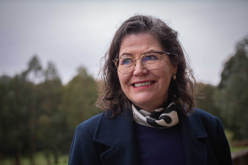 A woman with short brown hair, glasses, wearing a blue jacket and white scarf.