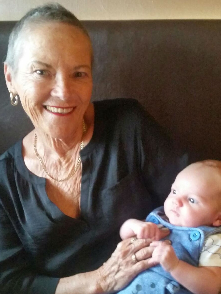 An older woman with very short grey hair holds a baby and smiles at the camera