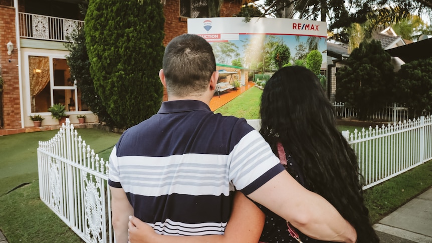 A man and woman hugging in front of a for sale sign at a house