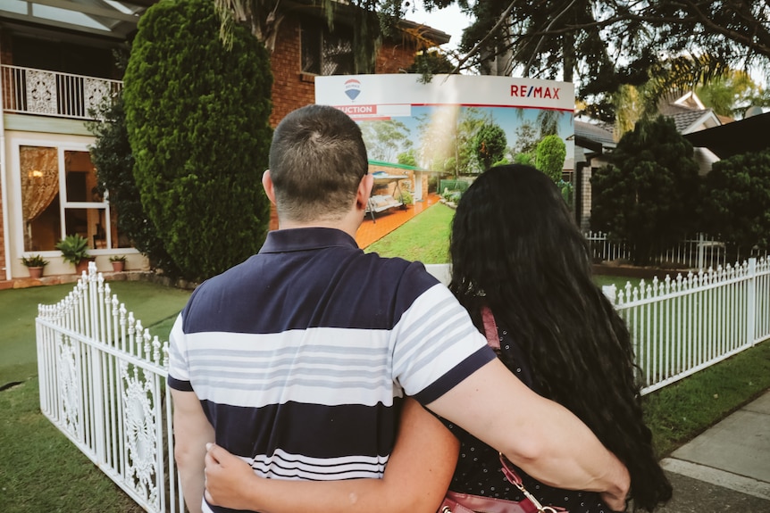 A man and a woman hugging in front of a for sale sign in a house