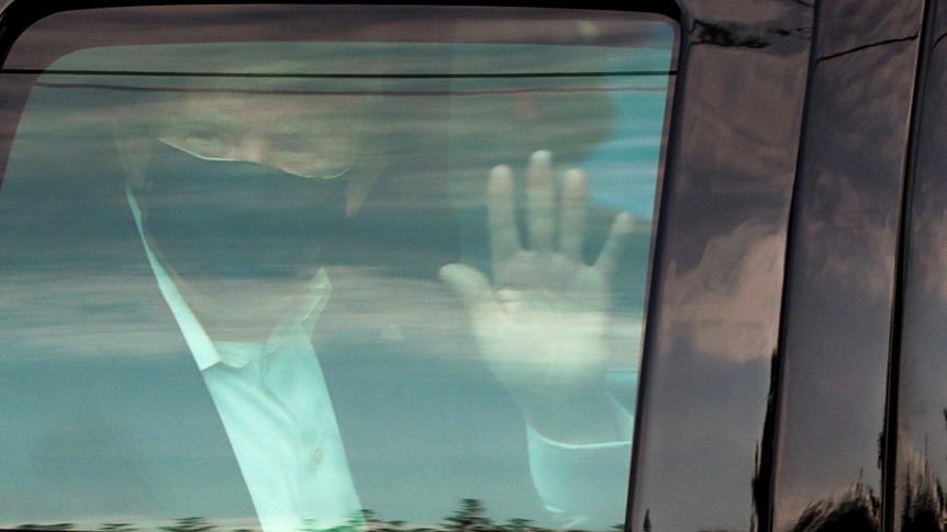 Donald Trump wearing a mask and waving seen therough the window of a car.