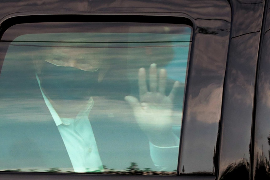 Donald Trump wearing a mask and waving seen therough the window of a car.