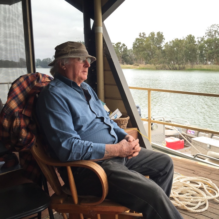 Rodney West sits on a rocking chair on the back deck of his houseboat and looks out at the river.