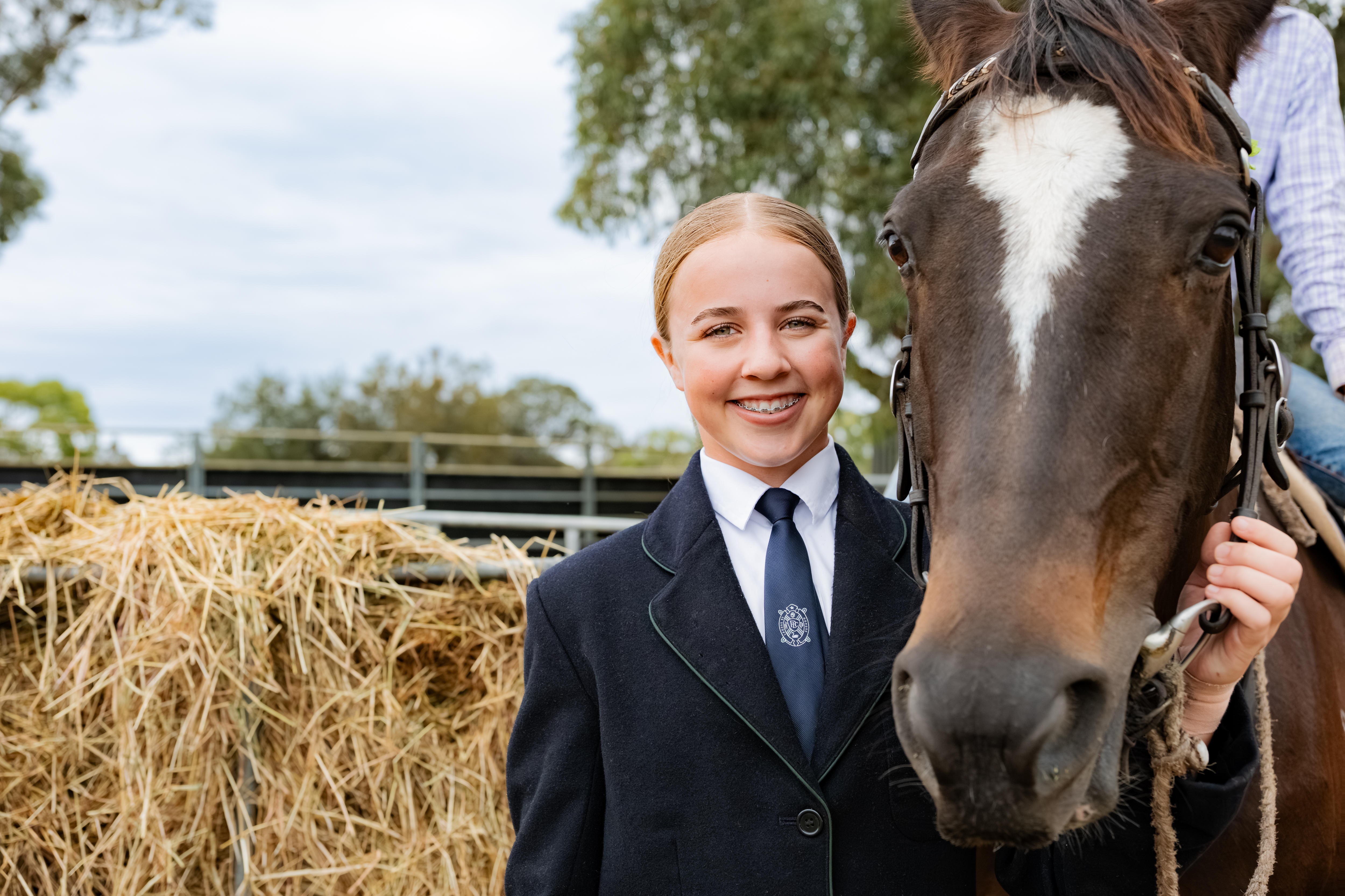 Una joven con uniforme escolar sonríe junto a un caballo en una propiedad rural.