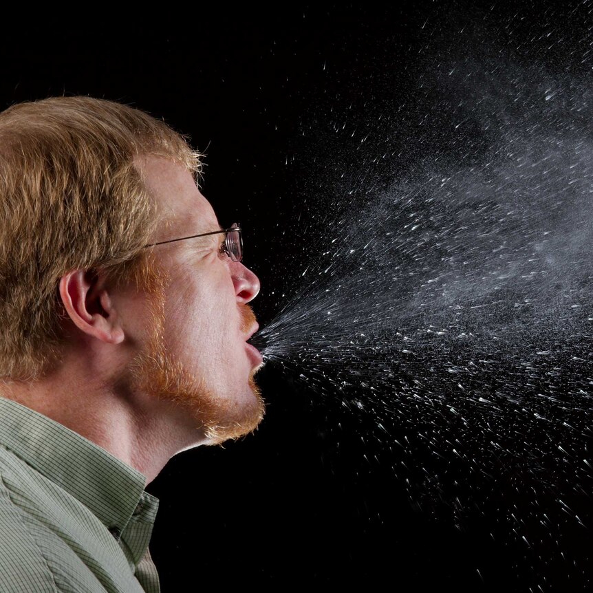 A man mid-sneeze against a black background, showing the salivary drops expelled from his mouth.