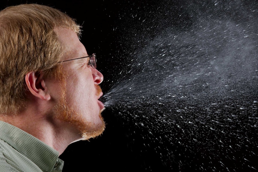 A man mid-sneeze against a black background, showing the salivary drops expelled from his mouth.