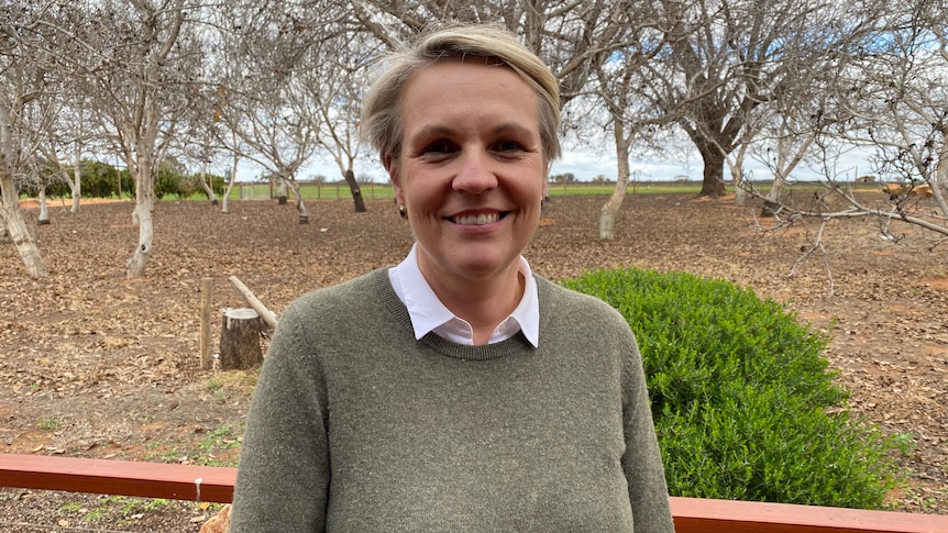 A woman with short hair wearing a grey jumper standing in front of trees. 