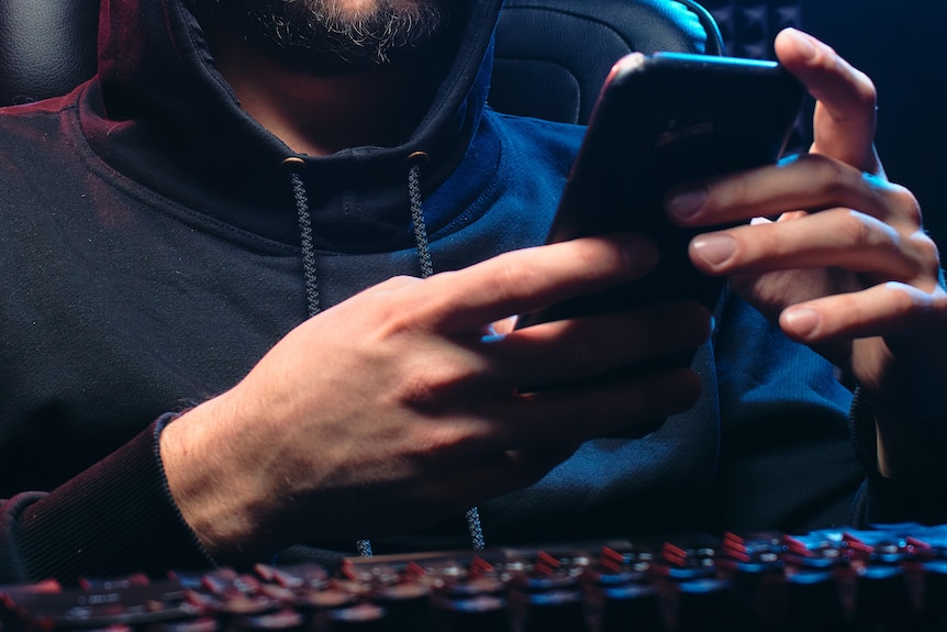 A bearded man in a hoodie operates a phone in an ill-lit room.