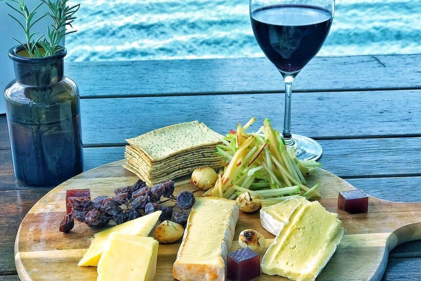 A platter of cheese, gourmet crackers, currants, quince paste cubes and a glass of red wine.