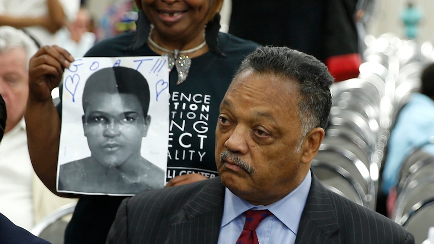 A woman holding a poster of Muhammad Ali stands behind Reverend Jesse Jackson.