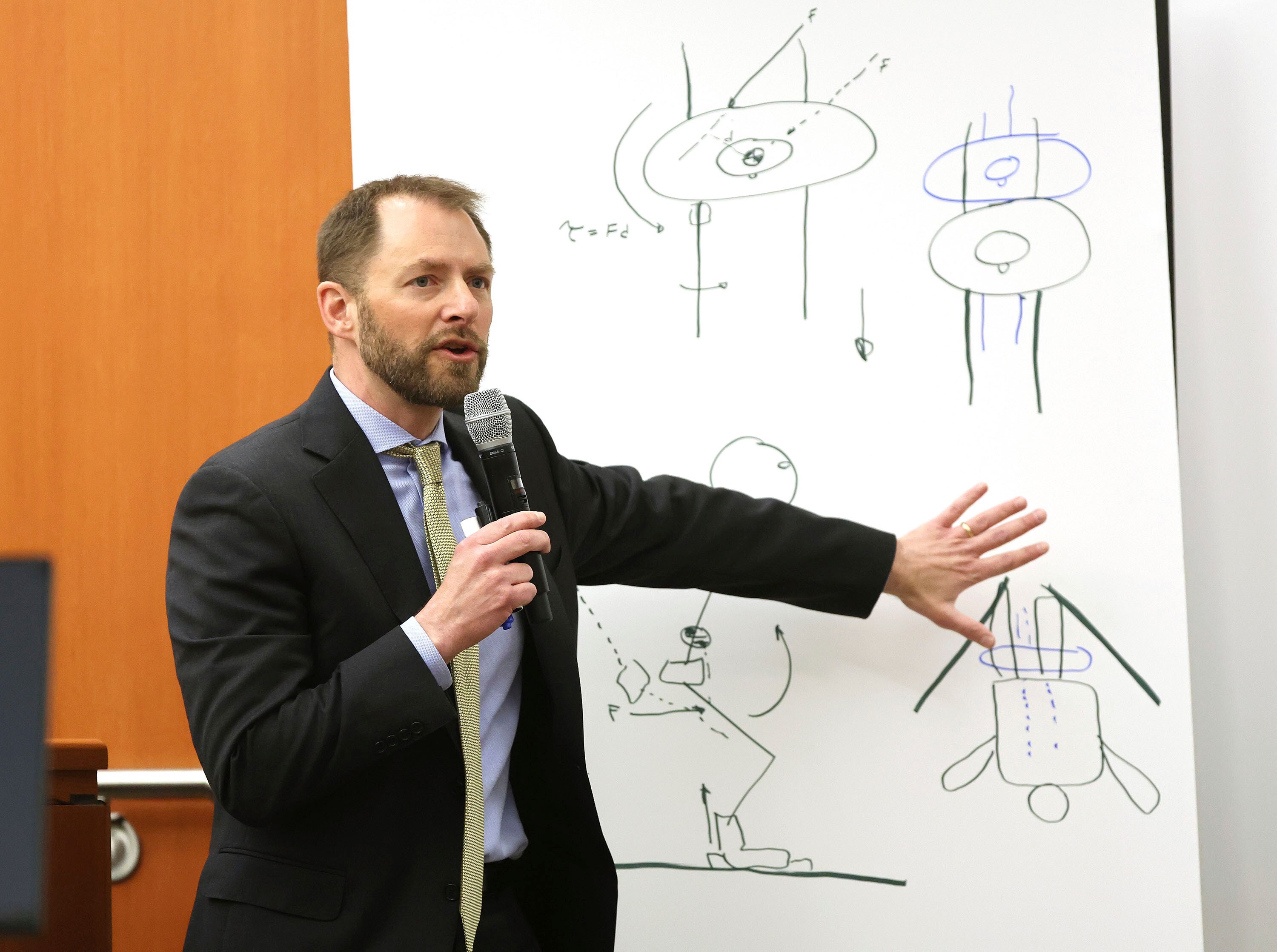 A man wearing a suit gestures at a whiteboard covered with simple drawings.