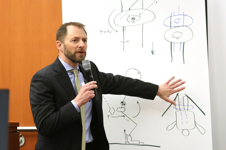 A man wearing a suit gestures at a whiteboard covered with simple drawings.