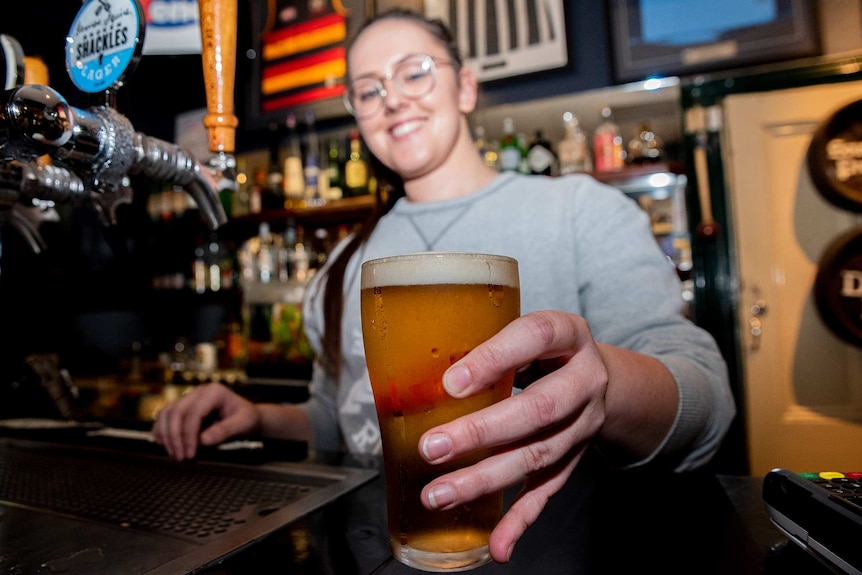 A woman holds out a freshly poured beer from behind a bar.