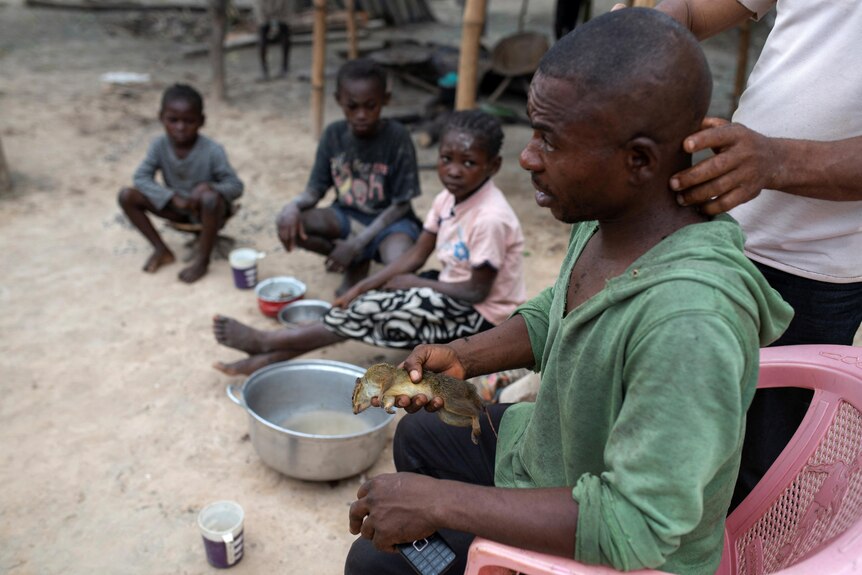 a man sits in a chair with a bush rat in his hand as someone checks his neck and his three children watch on