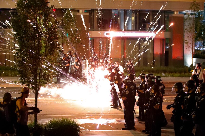 At night, a firework explodes by a police line amid a crowd of protesters in the middle of a street.