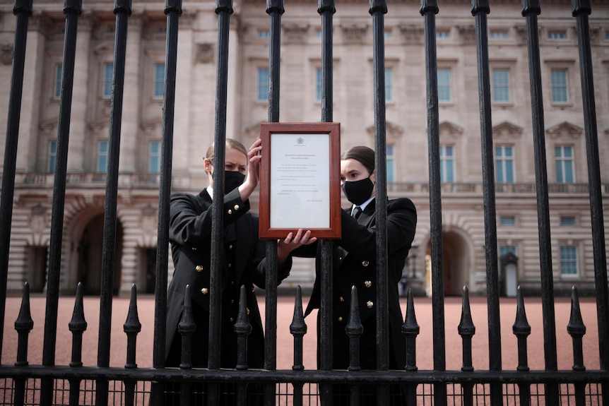 Two people attach a framed notice to a steel fence in front of Buckingham Palace.