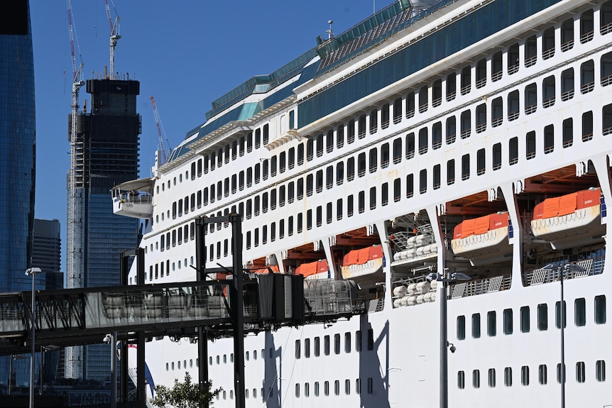 The side of a large white cruise ship with a skyscraper in the background.