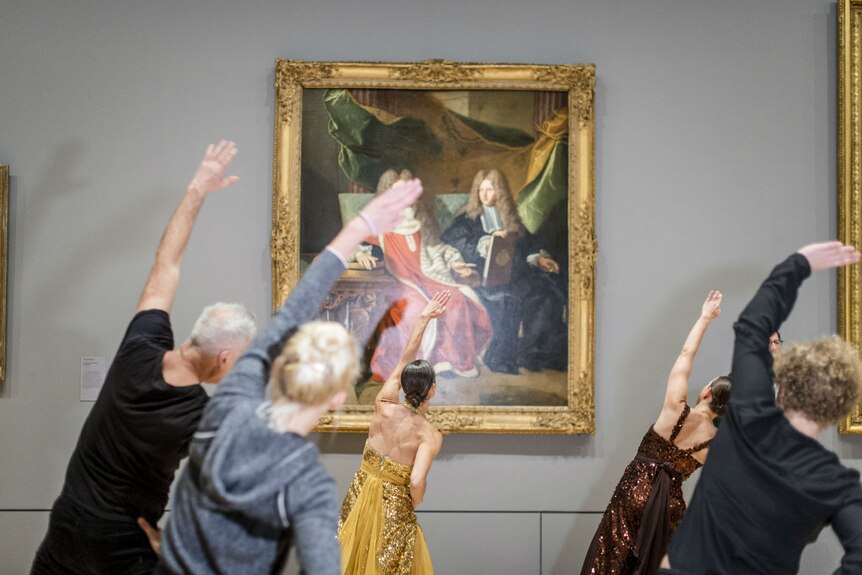 People stretch in front of a painting in a gold frame hung in a gallery.