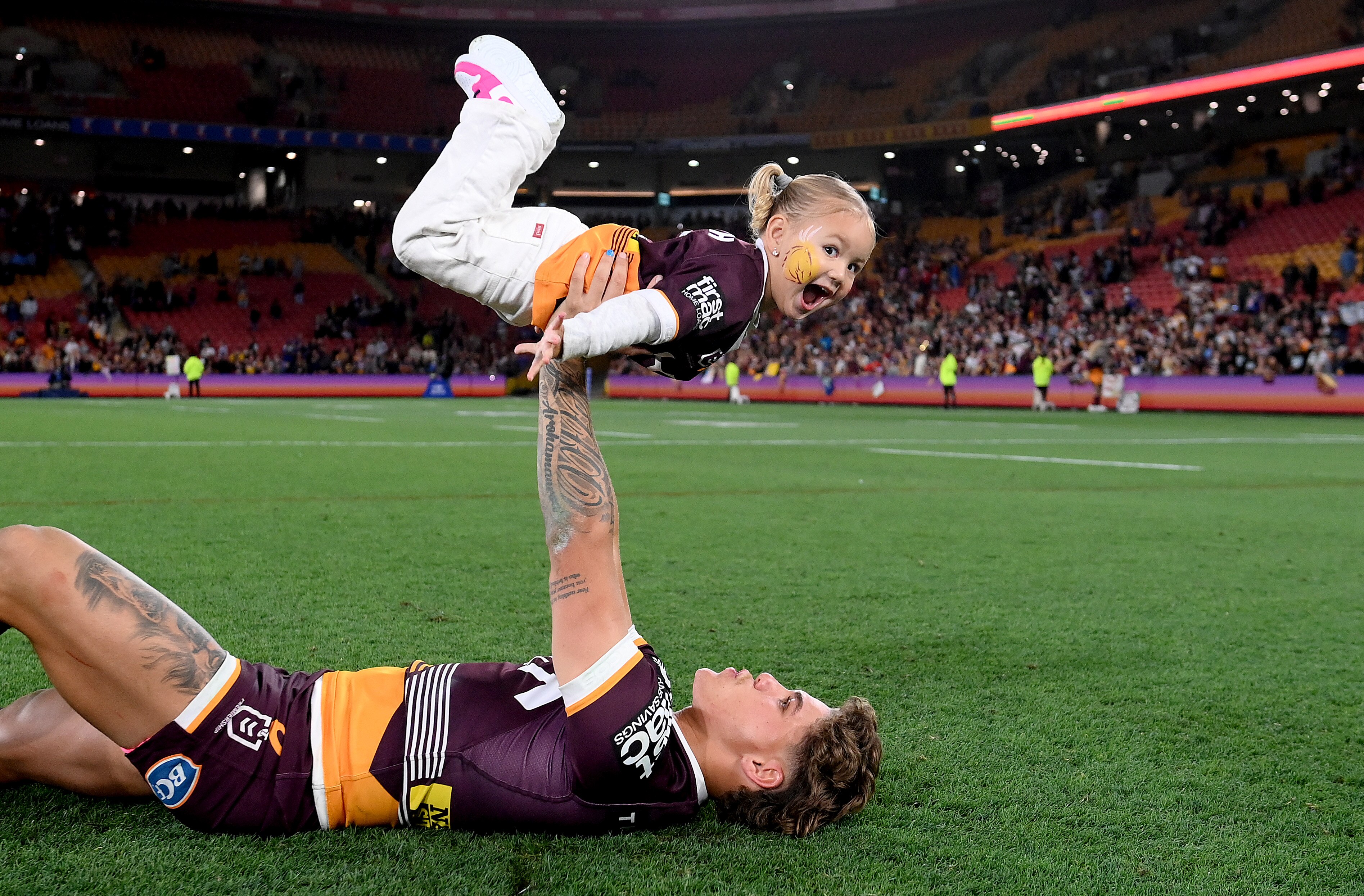 A Brisbane Broncos NRL player lies on the pitch at Lang Park holding his daughter up in the air in celebration.