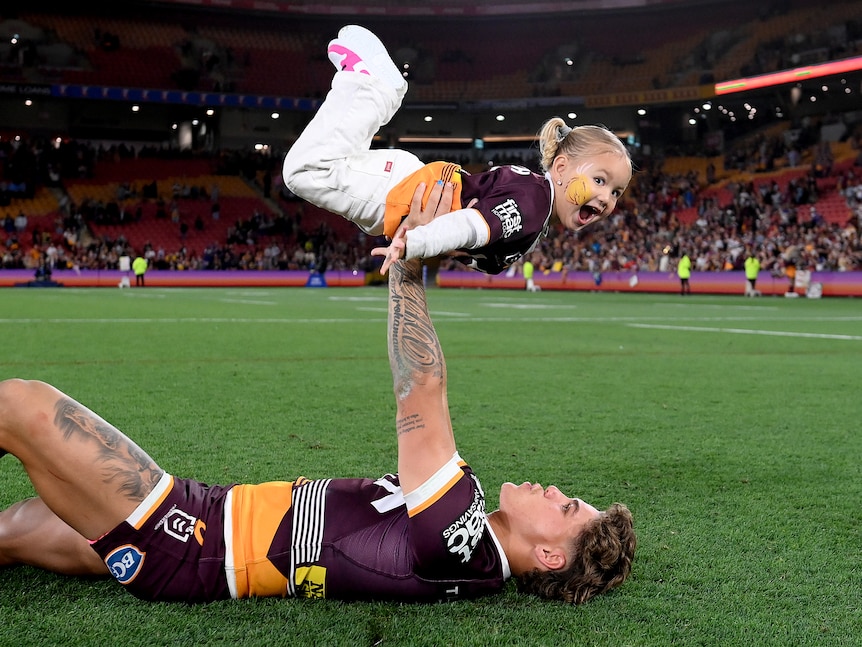 A Brisbane Broncos NRL player lies on the pitch at Lang Park holding his daughter up in the air in celebration.