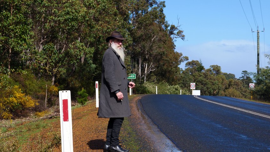 A man holding a cigarette hitchhiking along a highway in Greenbushes.