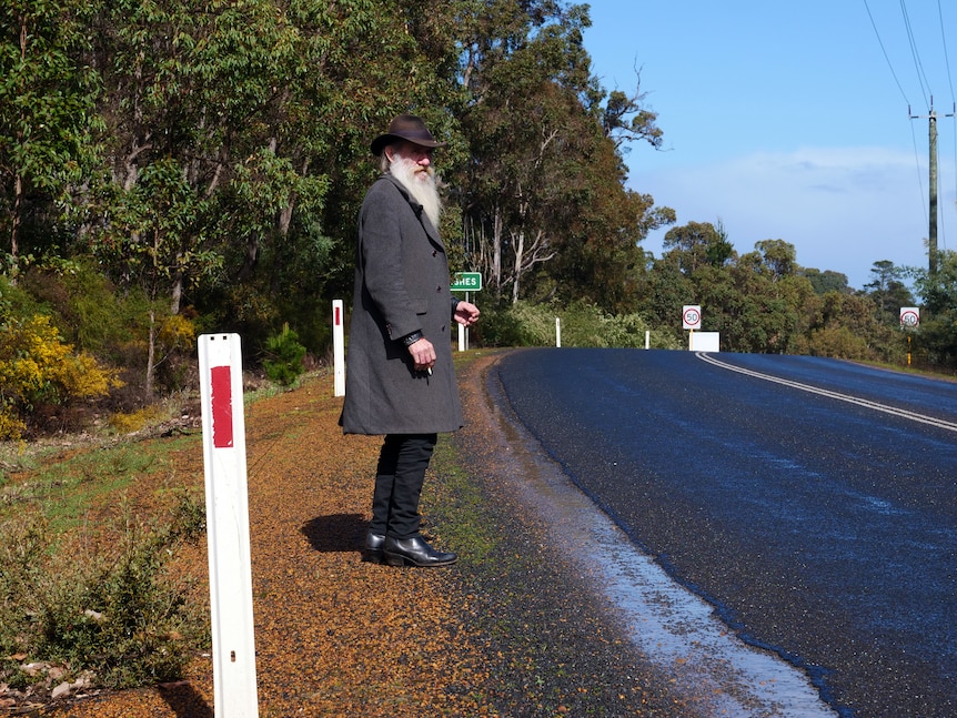 A man holding a cigarette hitchhiking along a highway in Greenbushes.