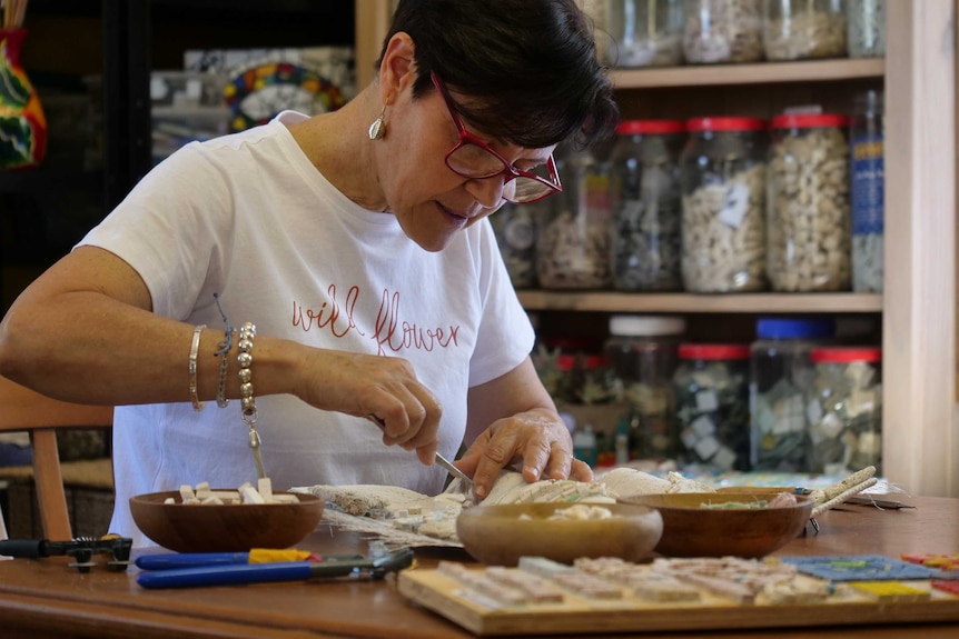 A woman with short dark hair, wearing glasses, sits at a wooden table creating a mosaic with an ocean theme.