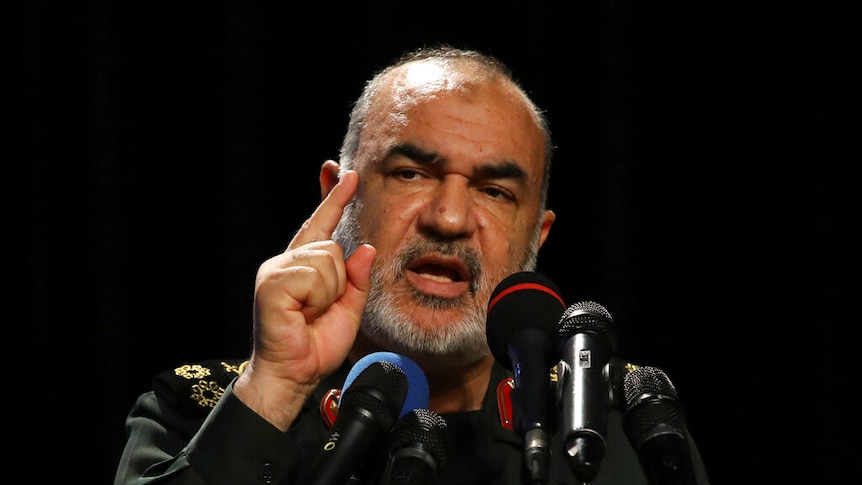Hossein Salami, in military uniform, gesticulates behind a string of multi-coloured microphones in front of a black backdrop.