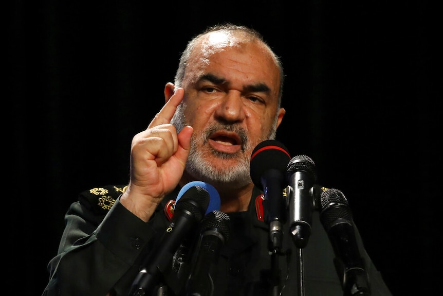 Hossein Salami, in military uniform, gesticulates behind a string of multi-coloured microphones in front of a black backdrop.