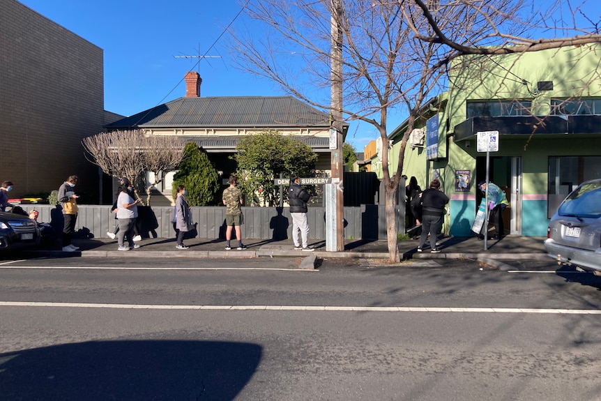 People stand in a queue to be tested for COVID-19 on a sunny suburban street.